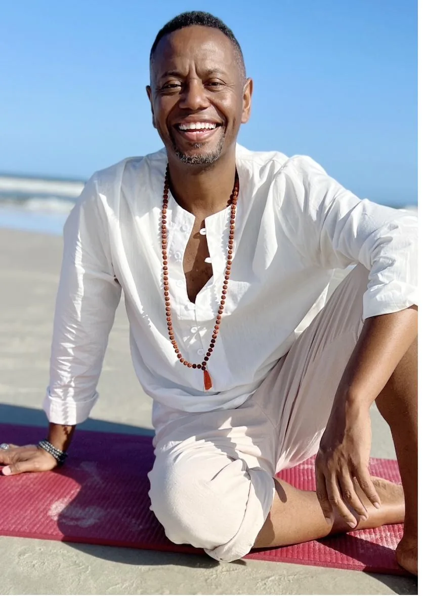 A.D. Roberts smiling on the beach in white, seated on a yoga mat